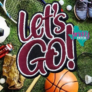 May include: A maroon and white embroidered patch featuring the text Let's Go! in a bold, script-style font with a thick black outline. The patch is layered over a background of various sports equipment including a ball, glove, and racket.