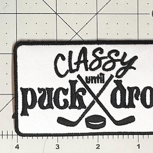 May include: White rectangular patch with black embroidered text and hockey graphics. The text reads "Classy until puck drop" with crossed hockey sticks and a puck. The patch is approximately 10 cm wide and 5 cm tall.