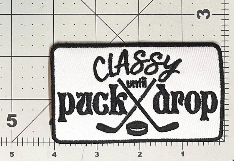 May include: White rectangular patch with black embroidered text and hockey graphics. The text reads "Classy until puck drop" with crossed hockey sticks and a puck. The patch is approximately 10 cm wide and 5 cm tall.