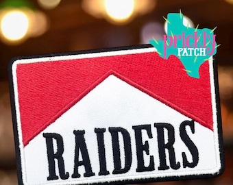 May include: A rectangular embroidered patch featuring a red and white chevron design with the word RAIDERS stitched in bold black lettering across the centre. The patch has a thick black border and a teal watermark logo in the top right corner.