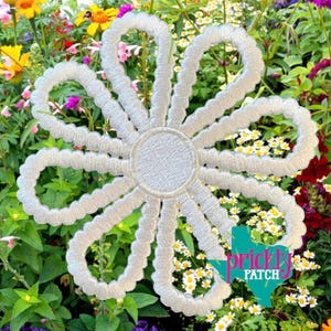 May include: White embroidered flower with eight looped petals and a circular center. The flower is set against a backdrop of colorful flowers and greenery. The image includes the text "Prickly Patch" in a Texas-shaped graphic.