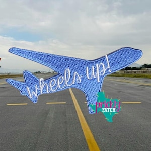 May include: A blue embroidered airplane patch with the words "wheels up!" in white script. The patch is in the shape of an airplane and is set against a blurred background of an airport runway and sky.
