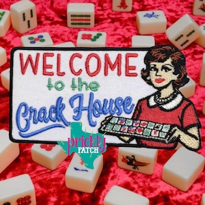May include: Embroidered patch with the words "WELCOME to the Crack House" in red and blue. A woman in a red dress and pearl necklace holds a Mahjong tile tray. Mahjong tiles are scattered on a red velvet surface.