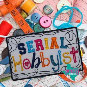 May include: Embroidered patch with the words "SERIAL HOBBYIST" in colorful letters. The patch is surrounded by a black border and sits on a background of sewing notions, including buttons, thread spools, and a measuring tape.