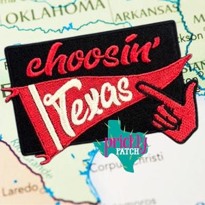 May include: A rectangular black embroidered patch featuring the red text choosin' above a red pennant flag with the white text Texas. A red hand with an extended index finger points toward the right side of the patch design.