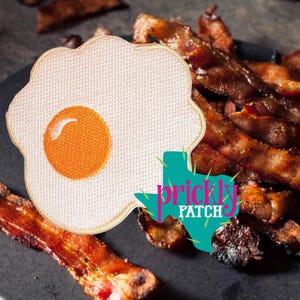 May include: Embroidered patch featuring a fried egg design with a white background and a yellow yolk. The patch is next to strips of cooked bacon. The text "prickly patch" is in the image.