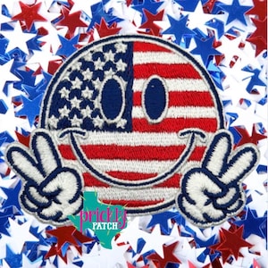 May include: Embroidered patch featuring a smiley face design with the American flag on one side and peace signs. The smiley face has a red, white, and blue color scheme. The background is filled with red, white, and blue star-shaped confetti.