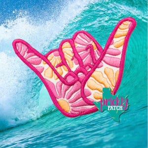 May include: An embroidered shaka hand sign patch featuring a vibrant floral pattern in shades of pink, orange, and white with a bright pink stitched border. A teal-colored Texas-shaped logo with the text Prickly Patch is visible at the bottom.