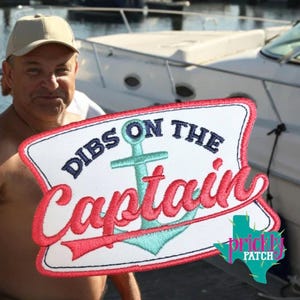 May include: Embroidered patch with the text "Dibs on the Captain" in red and navy blue, featuring a teal anchor design. The patch has a white background and a red border. The patch is in front of a boat.