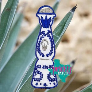 May include: Embroidered patch in the shape of a tequila bottle, featuring a white base with blue and tan detailing. The design includes floral and geometric patterns, with the text "prickly patch" in a turquoise color.