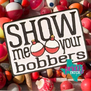 May include: A rectangular patch with the text "SHOW me your bobbers" in black lettering, featuring two bobber illustrations. The patch is surrounded by various red, white, and orange fishing bobbers.