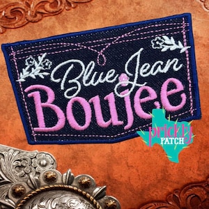 May include: Embroidered patch with the words "Blue Jean Boujee" in white and pink script. The patch is rectangular with a dark blue background and a blue border. Floral accents and a Texas-shaped logo are also present.