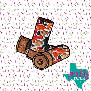 May include: Embroidered patch featuring two shotgun shells. The shells have a camouflage pattern in shades of orange, brown, and black. The patch has a brown border and a logo in the bottom right corner.