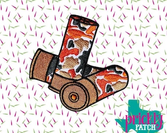 Puede incluir: Parche bordado con dos cartuchos de escopeta. Los cartuchos tienen un patrón de camuflaje en tonos naranja, marrón y negro. El parche tiene un borde marrón y un logotipo en la esquina inferior derecha.