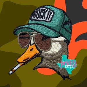May include: Embroidered patch featuring a duck wearing sunglasses and a green cap with the text "BUCK IT". The duck has a cigarette in its beak. The background is camouflage and orange.