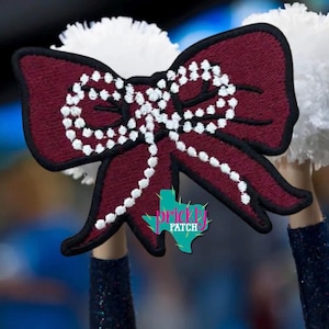 May include: A maroon embroidered bow patch with a white beaded-style outline and center detail. The patch features a black border and is layered over white pom-poms. A teal Texas-shaped logo with the text Prickly Patch is visible on the bow.