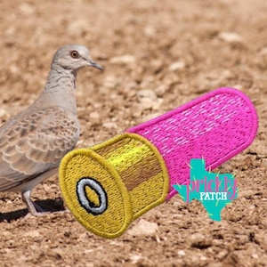 May include: An embroidered patch shaped like a shotgun shell with a bright pink body and a gold metallic-colored base featuring a central silver primer. A teal-colored graphic shaped like the state of Texas with the text Prickly Patch is visible.