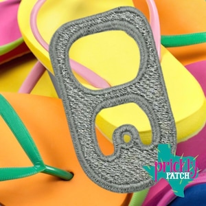 May include: A silver-coloured embroidered patch shaped like a soda can pull tab. The patch features a metallic grey thread texture with a distinct cutout design. It is positioned over a background of colourful yellow, orange, and pink flip-flops.