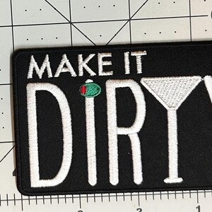 K&ouml;nnte beinhalten: Schwarzer rechteckiger Aufn&auml;her mit wei&szlig; gesticktem Text &bdquo;MAKE IT DIRTY&ldquo;. Eine gr&uuml;ne Olive mit roten Akzenten ist &uuml;ber dem Buchstaben &bdquo;R&ldquo; gestickt. Rechts ist ein wei&szlig; gesticktes Martiniglas.