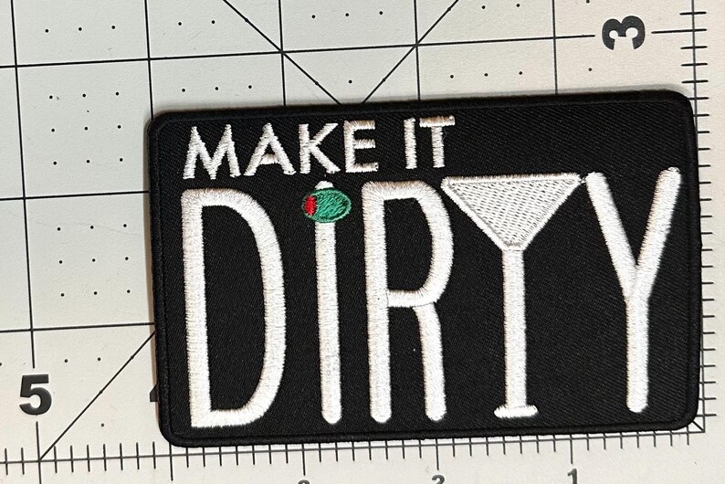 K&ouml;nnte beinhalten: Schwarzer rechteckiger Aufn&auml;her mit wei&szlig; gesticktem Text &bdquo;MAKE IT DIRTY&ldquo;. Eine gr&uuml;ne Olive mit roten Akzenten ist &uuml;ber dem Buchstaben &bdquo;R&ldquo; gestickt. Rechts ist ein wei&szlig; gesticktes Martiniglas.