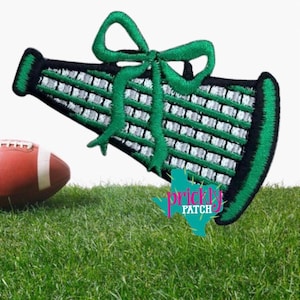 May include: An embroidered patch shaped like a megaphone with a green bow on top. The megaphone features black and silver striped detailing. A teal logo with the text "prickly PATCH" is visible in the lower right corner of the patch design.