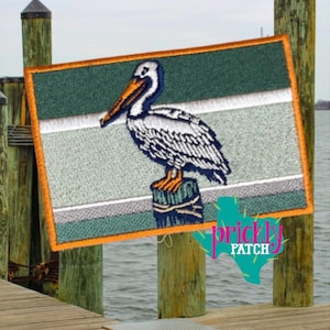 May include: Embroidered patch featuring a pelican perched on a wooden piling. The patch has a rectangular shape with a green, gray, and white striped background and an orange border. The text "prickly patch" is visible.