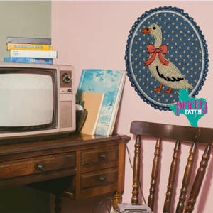 May include: Embroidered patch featuring a white goose with a red bow, set against a blue polka-dot background. The patch is framed by a scalloped border. The patch is affixed to a pink wall, near a vintage television and wooden desk.