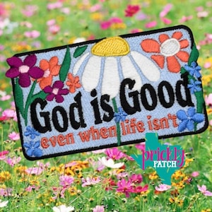 May include: Embroidered patch with the words "God is Good even when life isn't" in black and orange. The patch features a light blue background with embroidered flowers in shades of purple, orange, white, and blue. The patch has a black border.