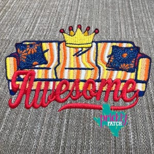 May include: Embroidered patch featuring a cartoon couch with a yellow crown. The couch has orange, yellow, and white stripes, with blue pillows. The word "Awesome" is in red script. The patch has a gray background.