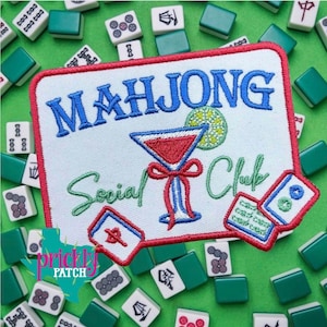 May include: Embroidered patch with the words "MAHJONG Social Club" in blue and green. The patch features a red and blue cocktail glass design and is surrounded by red stitching. Mahjong tiles are scattered around the patch on a green surface.
