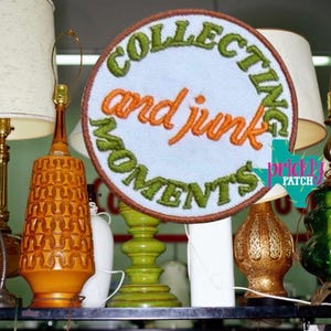 May include: A round patch with the words "COLLECTING and junk MOMENTS" embroidered in green and orange thread. The patch is surrounded by a brown border and set against a light blue background. The patch is displayed with several vintage lamps.