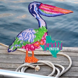 May include: A colorful embroidered patch of a pelican with a long, rainbow-colored beak. The bird has blue, green, pink, and orange details. The patch is on a wooden dock with a rope and the text "Prickly Patch".