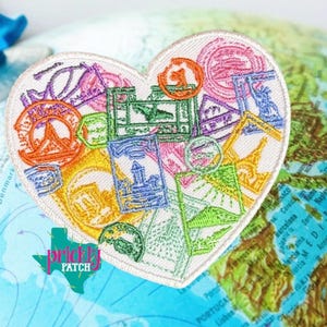 May include: Heart-shaped embroidered patch featuring colorful travel stamps. The patch has a white border and includes stamps in various shapes and colors, including orange, green, and blue. The patch is placed on a map.