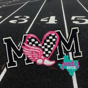 May include: Embroidered patch featuring the letters "MOM" in black with white trim. A pink heart with a checkered pattern and wings is centered between the letters. The patch is on a black track surface with white lane lines and numbers.