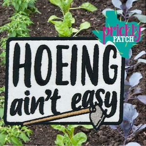 May include: Embroidered patch with the text "HOEING ain't easy" in black on a white background, featuring a small hoe illustration. The patch is set against a backdrop of a garden with green plants and soil.