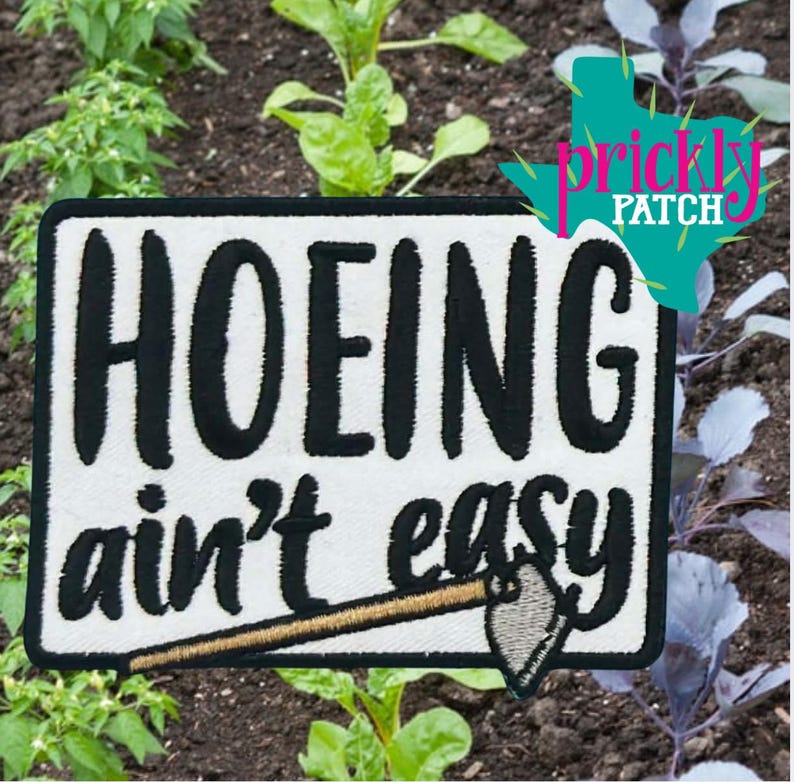 May include: Embroidered patch with the text "HOEING ain't easy" in black on a white background, featuring a small hoe illustration. The patch is set against a backdrop of a garden with green plants and soil.