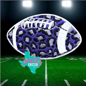 May include: An embroidered patch shaped like an American football, featuring a purple and grey leopard print pattern with white stitched laces and white stripes at each end, finished with a white border edge around the entire oval shape.