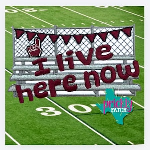 May include: An embroidered patch featuring a football stadium bleacher design with a maroon pennant banner, a foam finger icon, and the text "I live here now" in maroon thread. The design includes a white chain-link fence and a green field background.