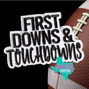 May include: A white embroidered patch with black lettering that reads FIRST DOWNS & TOUCHDOWNS, positioned over a brown and white football. The patch features a clean, bold font style with a distinct white border surrounding the black text.