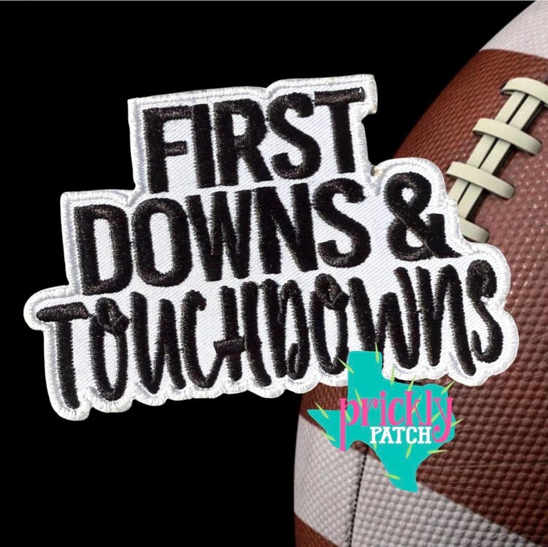 Puede incluir: Un parche bordado blanco con letras negras que dice FIRST DOWNS & TOUCHDOWNS, colocado sobre un bal&oacute;n de f&uacute;tbol americano marr&oacute;n y blanco. El parche presenta un estilo de fuente negrita y limpia con un borde blanco distintivo alrededor del texto.