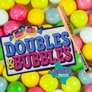 May include: An embroidered patch with the text "DOUBLES & BUBBLES" in blue and pink, a baseball bat, and a backdrop of colourful gumballs. The patch has a white border and a cartoon candy design at the top.