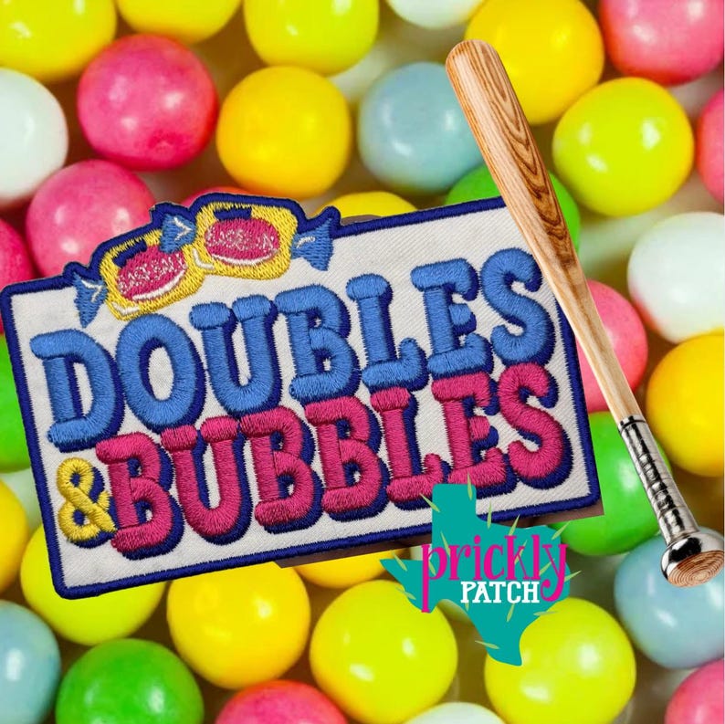 May include: An embroidered patch with the text "DOUBLES & BUBBLES" in blue and pink, a baseball bat, and a backdrop of colourful gumballs. The patch has a white border and a cartoon candy design at the top.