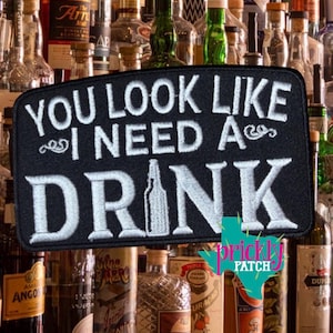 May include: Black rectangular patch with white embroidered text: "YOU LOOK LIKE I NEED A DRINK." A bottle graphic is below the text. The background features various liquor bottles.