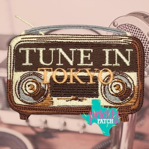 May include: Embroidered patch featuring a vintage radio design in shades of brown, beige, and gray. The text "TUNE IN TOKYO" is prominently displayed. The patch includes a silver antenna and a turquoise Texas-shaped logo.