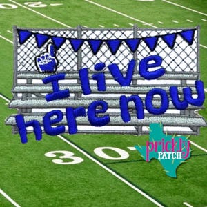 May include: An embroidered patch featuring a blue foam finger icon and the text "I live here now" in blue script lettering. The design includes a blue triangular pennant banner draped over a gray stadium bleacher graphic with a chain-link fence.