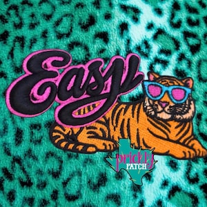May include: Embroidered patch featuring a tiger wearing blue sunglasses, with the word "Easy" in black and pink lettering. The patch is set against a teal leopard print background. A Texas-shaped logo is also visible.