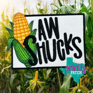 May include: Embroidered patch featuring a vibrant ear of corn with green husks and the text "AW SHUCKS" in black lettering on a white background. The patch includes a Texas-shaped logo with the words "prickly PATCH".