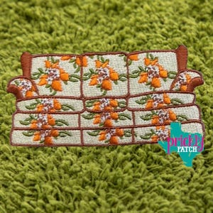 May include: Embroidered patch of a retro-style sofa with orange floral patterns on a cream background. The sofa has brown trim and armrests. The patch has the text "brickly PATCH" in a teal color.