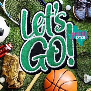 May include: An embroidered patch featuring the text "let's go!" in bright green thread with a thick white border on a black background. The lettering is in a bold, cursive script style, creating a high-contrast design for sports gear.