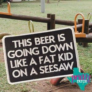 May include: A rectangular black embroidered patch with a white border and white stitched text that reads: THIS BEER IS GOING DOWN LIKE A FAT KID ON A SEESAW. The patch features a textured fabric surface and is displayed against a playground background.